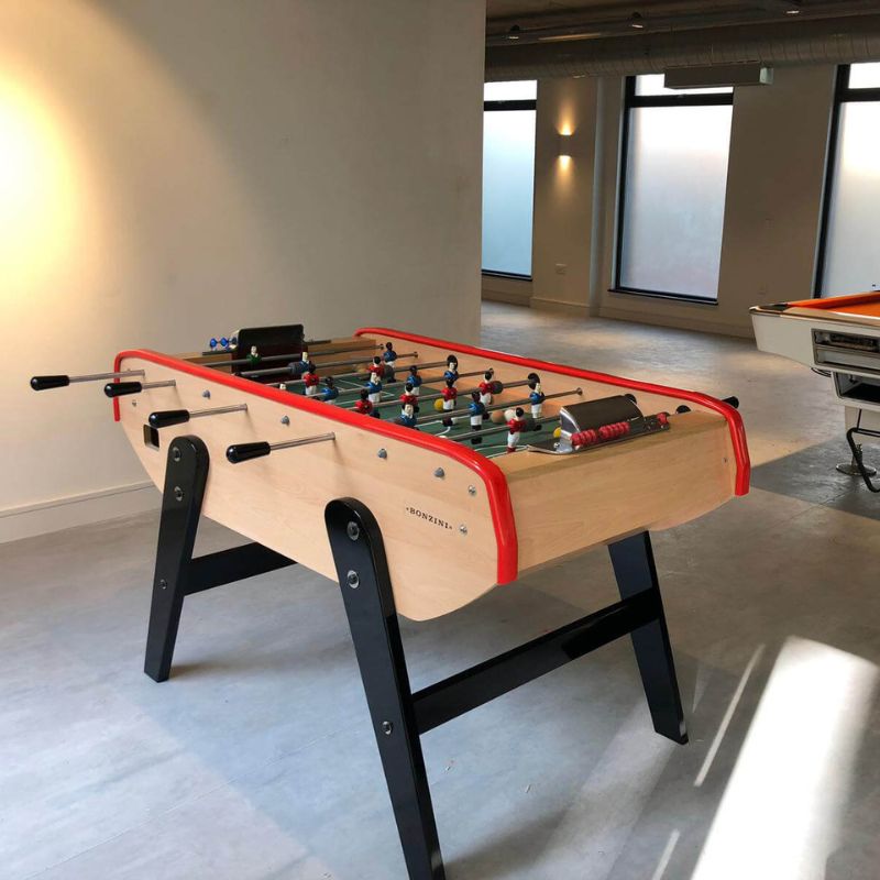 Modern foosball table with red trim in a bright, spacious game room with large windows.
