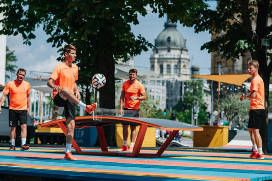 Four men playing Teqball outdoors on a curved table, wearing orange shirts and black shorts.