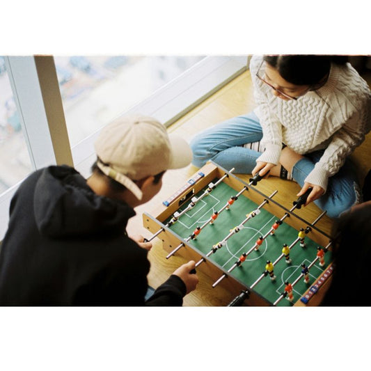Two people playing tabletop foosball by a window on wooden floor